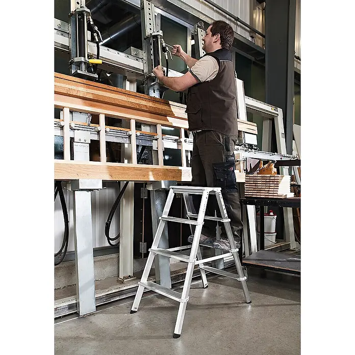 Munk Stehleiter Stufenstehleiter ML 2x6 Stufen - Arbeitshöhe: 2.950 m, Aluminium - Silberne Aluminium-Klappleiter, von einem Mann in einer Werkstatt für stabilen Zugang zu Maschinen verwendet.