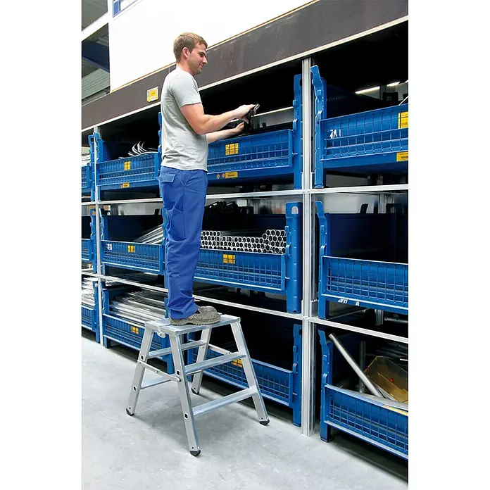 Munk Stehleiter Stufen Stehpodest klappbar, bedseitig begehbar 2x3 Stufen - Anzahl Tritte: 3 Stk., Arbeitshöhe: 2.690 m, Aluminium - Silberne Aluminium-Klapptritt, von einem Mann zum Erreichen hoher Regale in einem Lagerhaus genutzt.