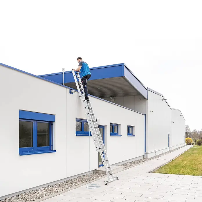 Munk Seilzugleiter Stufen- Seilzugleiter 2-teilig mit Handlauf und Ausstiegsholm 2x12 Stufen - Arbeitshöhe: 7.000 m, Belastbarkeit: 150 kg, Aluminium - Mann steigt auf einer Aluminium-Anlegeleiter auf das Dach eines modernen weißen Gebäudes.
