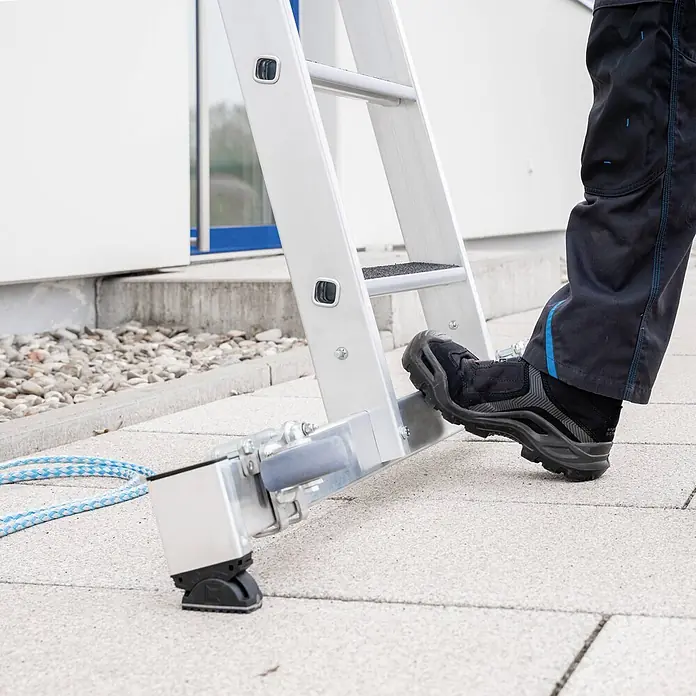 Munk Seilzugleiter Stufen- Seilzugleiter 2-teilig mit Handlauf und Ausstiegsholm 2x12 Stufen - Arbeitshöhe: 7.000 m, Belastbarkeit: 150 kg, Aluminium - Fuß eines Arbeiters auf der untersten Sprosse einer silbernen Aluminiumleiter mit verstellbarer Nivellierbasis.
