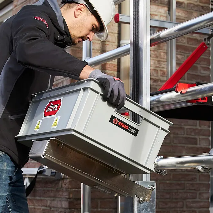 Altrex Rollgerüst Shuttle Carrier-Satz als Zubehör für Shuttle-Lift - Profil kurz ohne Befe -  - Mann in Schutzhelm lädt eine graue Altrex-Kunststoffkiste mit Ergo360-Logo auf eine Metallgerüstplattform.