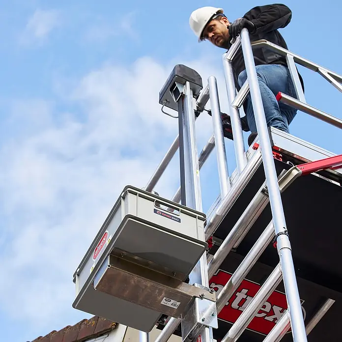 Altrex Rollgerüst Shuttle Carrier-Satz als Zubehör für Shuttle-Lift - Profil kurz ohne Befe -  - Altrex Arbeitsbühne mit grauer Werkzeugkiste, von einem Arbeiter in der Höhe unter blauem Himmel genutzt.