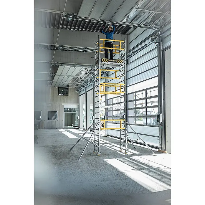 Munk Rollgerüst Flexxtower mit Standardgeländer bis 6,10m AH - Arbeitshöhe: 6.100 m - Fahrbares Gerüst, silber mit gelben Akzenten, wird von einer Person für Arbeiten an Deckeninstallationen verwendet.