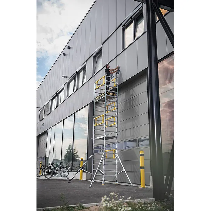Munk Rollgerüst Flexxtower mit Standardgeländer bis 6,10m AH - Arbeitshöhe: 6.100 m - Mobiles Aluminium-Rollgerüst mit gelben Sprossen und Geländern, verwendet von einem Mann an einer Gebäudefassade.