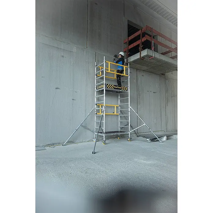 Munk Rollgerüst Flexxtower mit Standardgeländer bis 6,10m AH - Arbeitshöhe: 6.100 m - Aluminium-Rollgerüst mit gelben Akzenten. Ein Arbeiter nutzt ein Werkzeug auf der oberen Plattform in einem Betongebäude.
