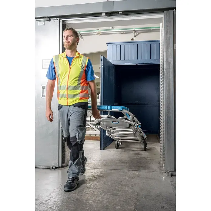 Zarges Teleskopleiter Teleskop Plattformleiter ZAP Masterflex Plus S gebördelt 3-5 Stufen - Anzahl Tritte: 5 Stk., Arbeitshöhe: 3.280 m, Belastbarkeit: 150 kg, Aluminium - Arbeiter transportiert eine zusammengeklappte, silberne Multifunktionsleiter mit blauen Akzenten und Rollen durch eine Industrietür.