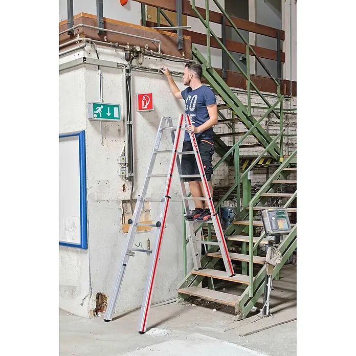 Hymer Teleskopleiter Treppenstehleiter Teleskopleiter mit patentierter Feststellvorrichtung 2x7 - Anzahl Tritte: 7 Stk., Arbeitshöhe: 3.300 m, Aluminium - Silberne Aluminium-Klappleiter mit roten Akzenten, von einem Mann zum Erreichen von Überkopfarbeiten in einem Industriegebäude verwendet.