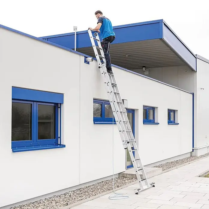 Munk Seilzugleiter Stufen- Seilzugleiter mit Nivello-Traverse und clip-step R13 2x12 Stufen - Anzahl Tritte: 12 Stk., Arbeitshöhe: 7.000 m, Aluminium - Mann benutzt eine silberne Anlegeleiter, um das Gebäudedach zu erreichen.