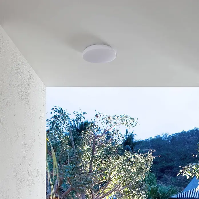 Lindby Außenwand- & Deckenleuchte Doki - weiß / opal, IP44 - Weiße, runde LED-Deckenleuchte an einer weißen Decke über einer Aussicht mit Bäumen.