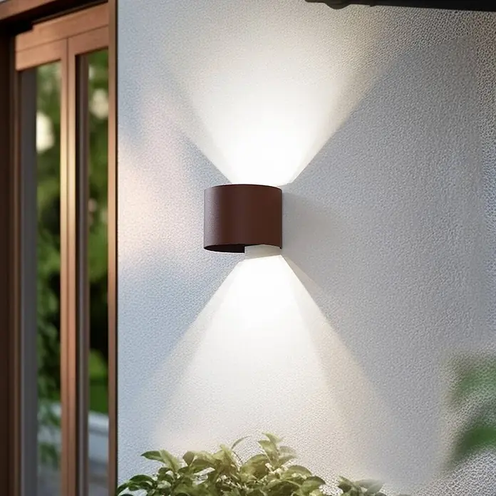 Lindby Außenwandleuchte Dara - braun / rost, IP54 - Zylindrische bronzefarbene Wandleuchte mit Up-Down-Beleuchtung auf einer strukturierten Wand neben einer Holztür und Pflanzen.