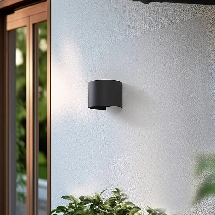 Lindby Außenwandleuchte Dara - alu / grau / zink, IP54 - Dunkelgraue, gekrümmte Wandleuchte an einer strukturierten Wand, neben einem Fenster mit Außengrün.