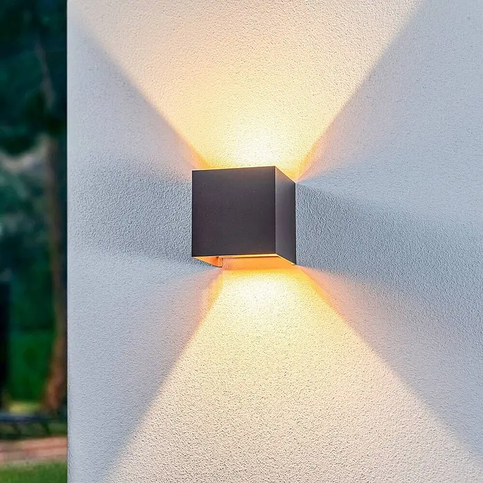 Lindby Außenwandleuchte Nivar - 10 x 10 x 10 cm, schwarz, IP54 - Dunkelgraue kubische Außenwandleuchte, die eine strukturierte Wand mit warmem Licht nach oben und unten beleuchtet.