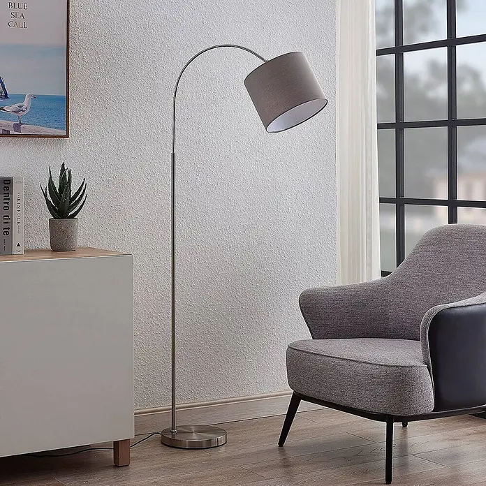 Lindby Stehleuchte Keriba - Höhe: 150,2 cm, alu / grau / zink, alu / grau / zink, E27 - Bogenförmige Stehlampe mit gebürstetem Metallsockel und grauem Stoffschirm, neben einem Sessel platziert.