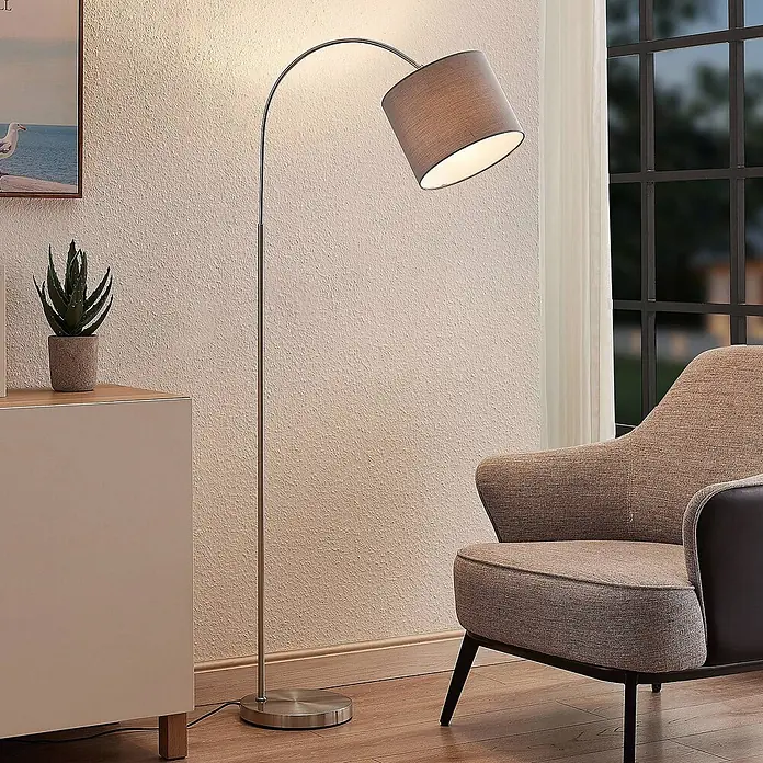 Lindby Stehleuchte Keriba - Höhe: 150,2 cm, alu / grau / zink, alu / grau / zink, E27 - Eine elegante Bogen-Stehleuchte mit silberner Oberfläche und grauem Stoffschirm, die einen gemütlichen Sessel in einem modernen Wohnzimmer beleuchtet.