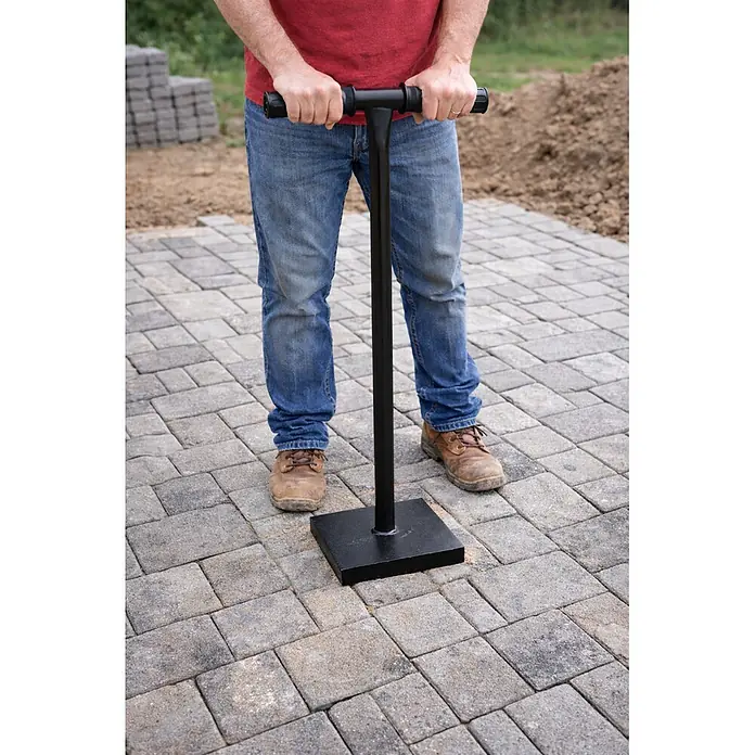 Küpper Betonstampfer - Stahl, 13 kg - Person benutzt schwarzes T-Griff-Pflasterwerkzeug mit quadratischem Sockel auf grauen Pflastersteinen.