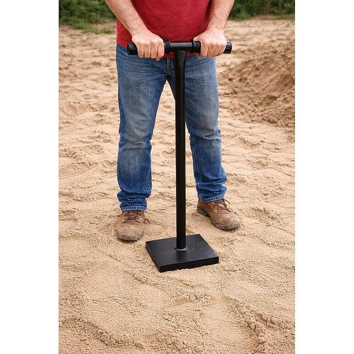Küpper Betonstampfer - Stahl, 13 kg - Person bedient einen schwarzen T-förmigen Sandstampfer mit quadratischer Basis auf Sand.