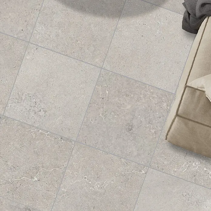 RAK Ceramics Feinsteinzeugfliese Valdi - 120 x 120 cm, Light Grey, Matt - Helle, graue, strukturierte Bodenfliesen mit Fugen, teilweise sichtbar ein beiges Sofa.