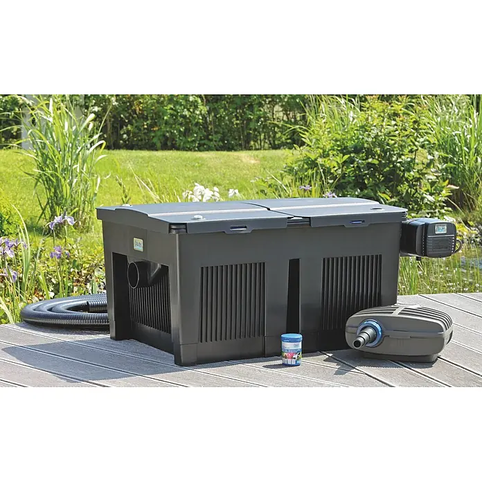 Oase BioSmart Durchlauffilter-Set 24000 - 24 W, Teichgröße: 24 m³, Fördermenge pro Std.: 5.300 l - Ein schwarzes Teichfiltersystem mit grauer Pumpe, Schlauch und Teichpflegebehälter steht auf einem Holzdeck neben einem Gartenteich.