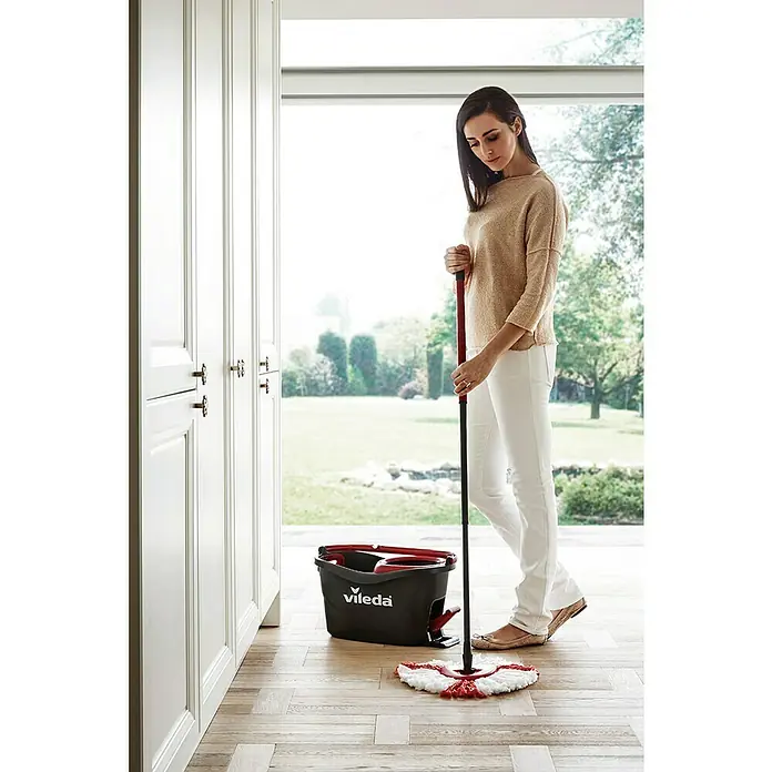 Vileda Set de mopa Turbo set - 3 pzs. - Mujer está limpiando un piso de madera con un juego de trapeador Vileda, con balde. El juego incluye un trapeador con mango telescópico y un balde negro con exprimidor rojo.