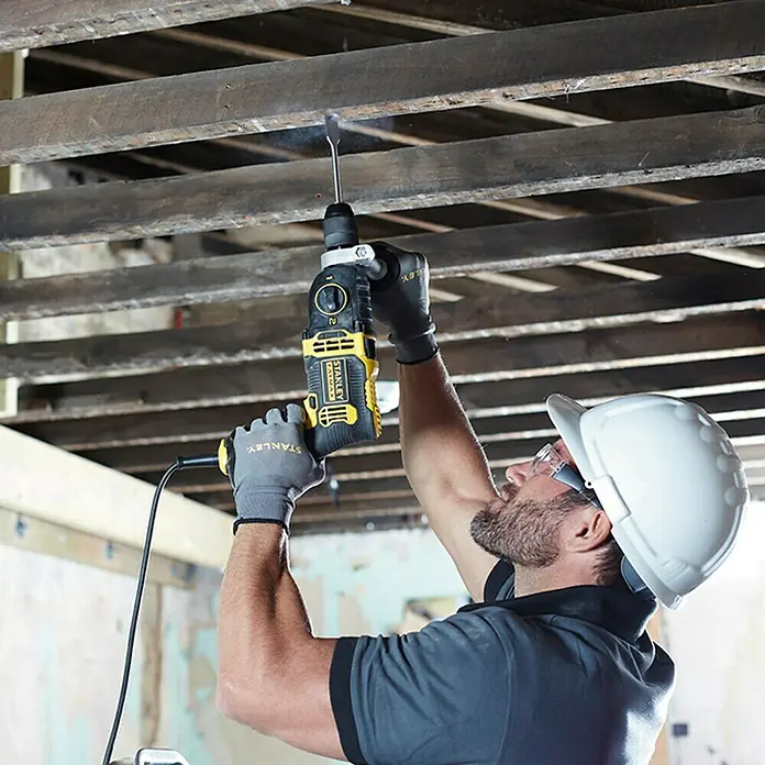 Stanley FatMax Taladro percutor FMEH850K-QS - 850 W, Portabrocas de sujeción rápida - Un trabajador de la construcción con casco está usando un taladro percutor SDS Stanley FATMAX para perforar un techo.