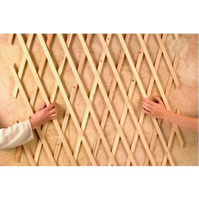 Nortene Celosía Trelliwood - An x Al: 0,5 x 1,5 m, Extensible, Natural - Aquí hay una valla de jardín hecha de listones de madera que dos manos están uniendo a una pared. La cerca tiene un patrón de rombos.