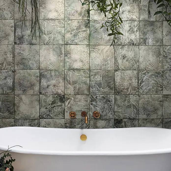 Feinsteinzeugfliese Amazonia - 24,8 x 24,8 cm, Grün, Matt - Weiße Badewanne und Messingarmaturen in einem Badezimmer mit botanischen Kachelmustern und hängendem Laub.