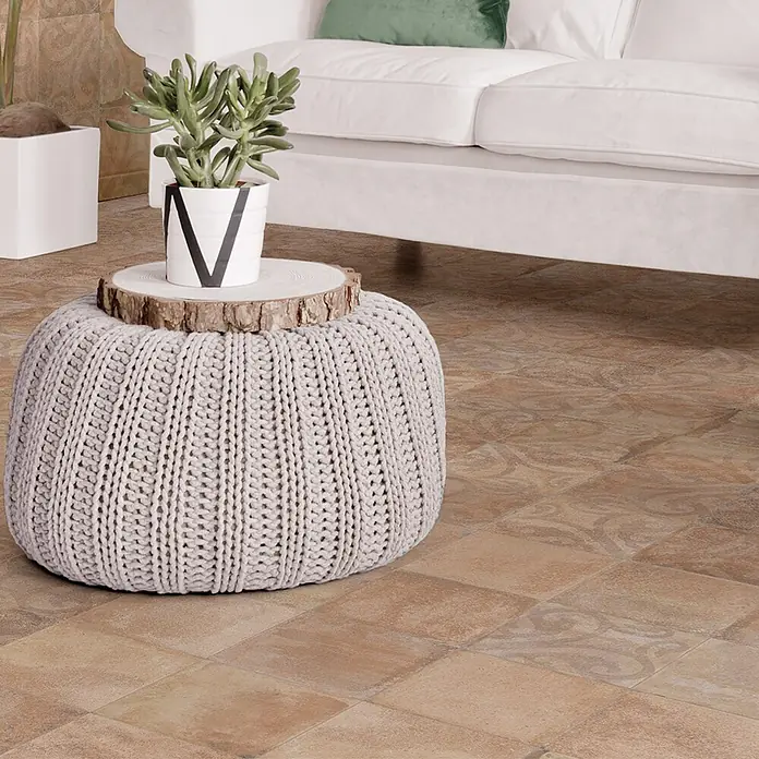Feinsteinzeugfliese Heredad Greek avena - 24,8 x 24,8 cm, Cotto, Matt - Hellgrauer Strickpouf, als Beistelltisch genutzt, mit einer Pflanze auf einer Holzscheibe, in einem Wohnzimmerambiente.