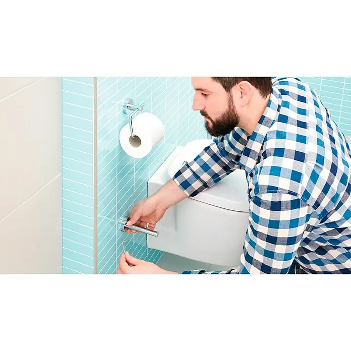 Tesa Smooz Portarrollos de repuesto - Cromo, Brillante - Un hombre en un baño está reparando un accesorio de baño con una herramienta, con una pared azul y un rollo de papel higiénico de fondo.