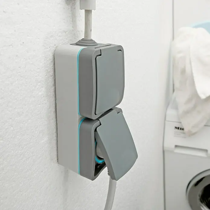 Voltomat Blueline Feuchtraum-Steckdose - 2-fach, Vertikal, IP54, Grau - Nahaufnahme einer Doppelsteckdose mit Klappdeckel an einer weißen Wand, ein Kabel ist eingesteckt.