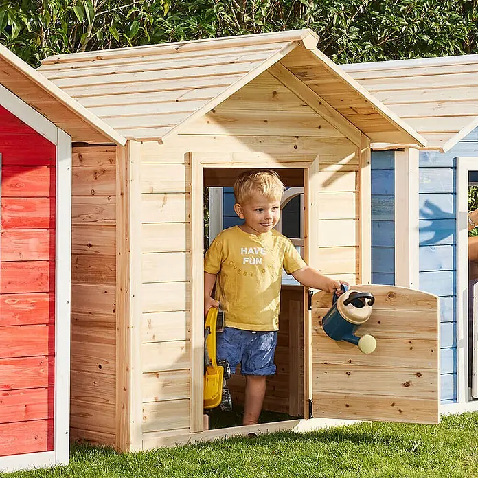Home Deluxe Spielhaus DER GROSSE PALAST - L x B x H: 111 x 118,2 x 132,5 cm, Holz, Natur - Ein kleines Kind spielt in einem Spielhaus aus Naturholz im Garten, hält einen Spielzeugbagger und eine Gießkanne.