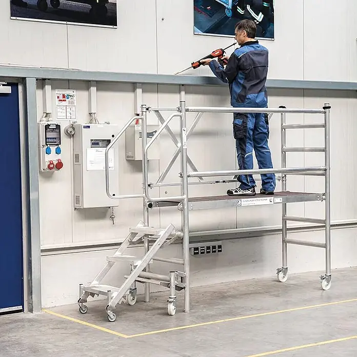 Munk Stehleiter Montageplattform mit Treppenzustieg 3m AH - Anzahl Tritte: 3 Stk., Arbeitshöhe: 3.000 m, Aluminium - Mobile Aluminium-Arbeitsplattform, auf der ein Arbeiter mit einem Werkzeug Arbeiten in der Höhe ausführt.