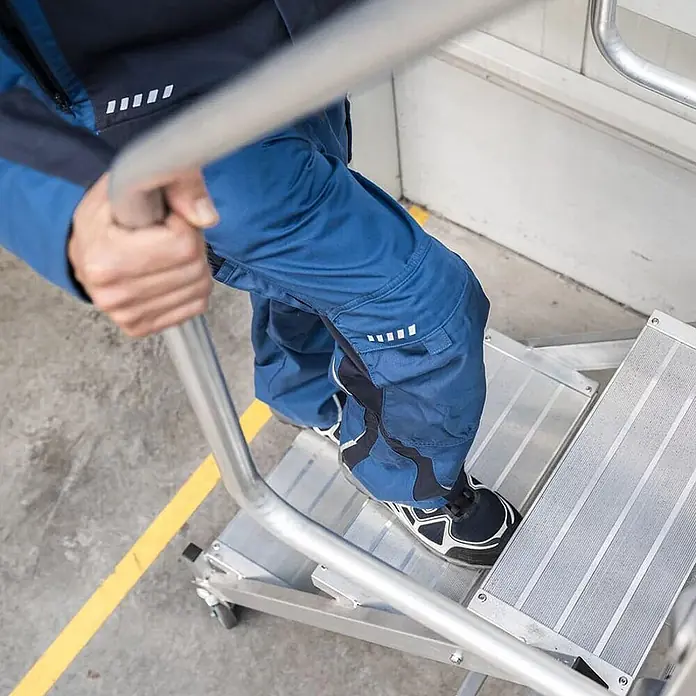 Munk Stehleiter Montageplattform mit Treppenzustieg 3m AH - Anzahl Tritte: 3 Stk., Arbeitshöhe: 3.000 m, Aluminium - Person nutzt einen mobilen Aluminium-Sicherheits-Klapptritt mit zwei rutschfesten Stufen und Handlauf.