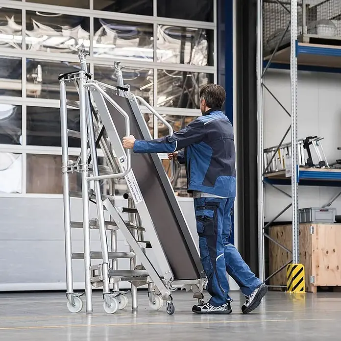 Munk Stehleiter Montageplattform mit Treppenzustieg 3m AH - Anzahl Tritte: 3 Stk., Arbeitshöhe: 3.000 m, Aluminium - Mobile Aluminium-Arbeitsplattform mit Stufen und schräger Fläche, von einer Person bewegt.