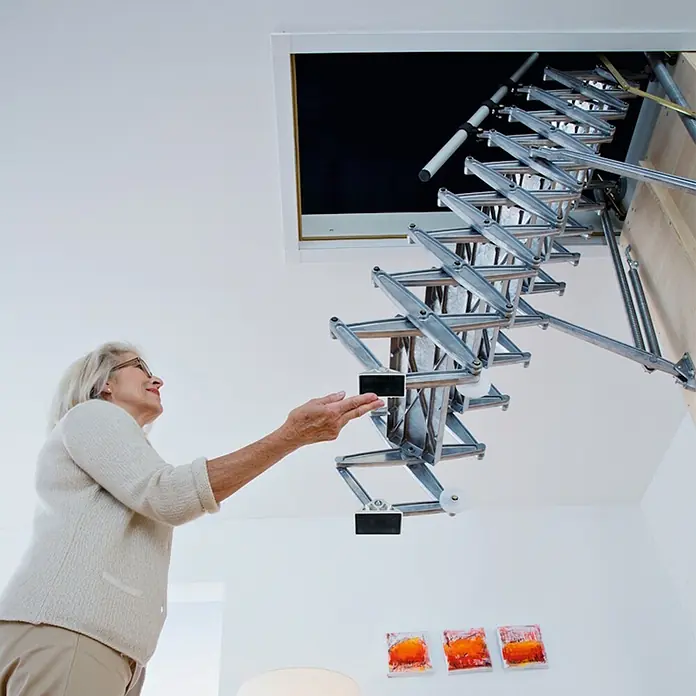 Roto Bodentreppe Scherentreppe Exklusiv 250cm max. Lichte Raumhöhe 120x60cm Rohbaumaß -  - Silberfarbene, ausziehbare Metall-Bodentreppe fährt aus einer Deckenöffnung herunter, eine Frau greift danach.
