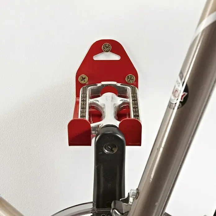 Mottez Gancho de pared para bicicletas B215V - Acero - Primer plano de un colgador de pedal de bicicleta rojo montado en la pared, que sujeta un pedal de bicicleta.
