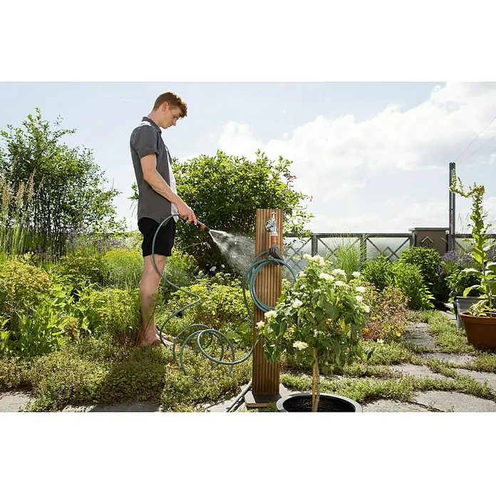 Kit de manguera en espiral City Gardening - Largo: 7,5 m - Un hombre riega un jardín exuberante con una manguera de jardín Gardena. La manguera está fijada a un poste de madera con grifo.