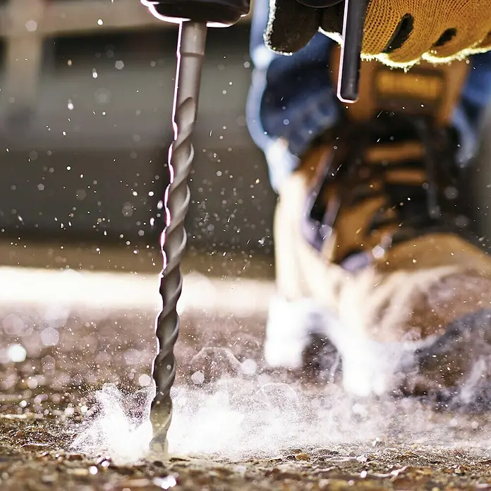 DeWalt Broca para hormigón y mampostería DT9502-QZ - 1 ud., Diámetro de la broca: 4 mm - Primer plano de una broca perforando una superficie, con escombros saliendo. La persona lleva guantes y botas.