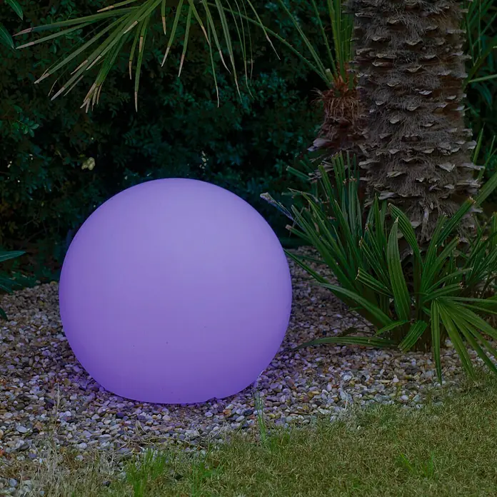 New Garden Bola solar LED Buly 30 Smart Tech - Altura: 26,5 cm, LED - Una esfera iluminada de color púrpura está en el jardín, rodeada de piedras, hierba y plantas verdes. La luz del jardín añade un toque de color.