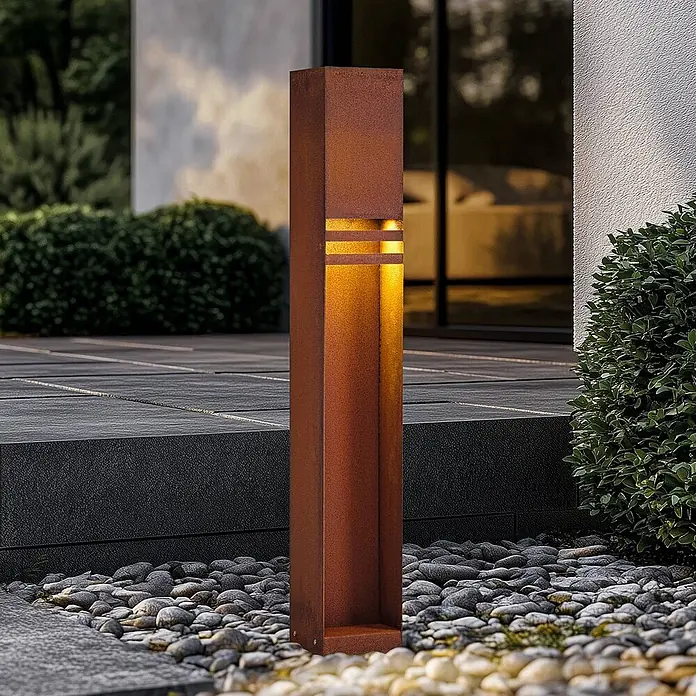Lucande Außenlaterne Oxido -  - Hohe, rechteckige Gartenleuchte in Rost-Optik (Cortenstahl), die warmes Licht durch horizontale Schlitze abgibt, in einem Kiesbeet.