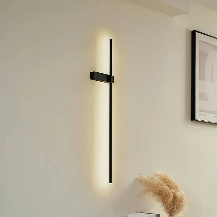 Lindby Wandleuchte Natsumi - schwarz - Moderne schwarze lineare Wandleuchte, die warmes Umgebungslicht abgibt, an einer cremefarbenen Wand.