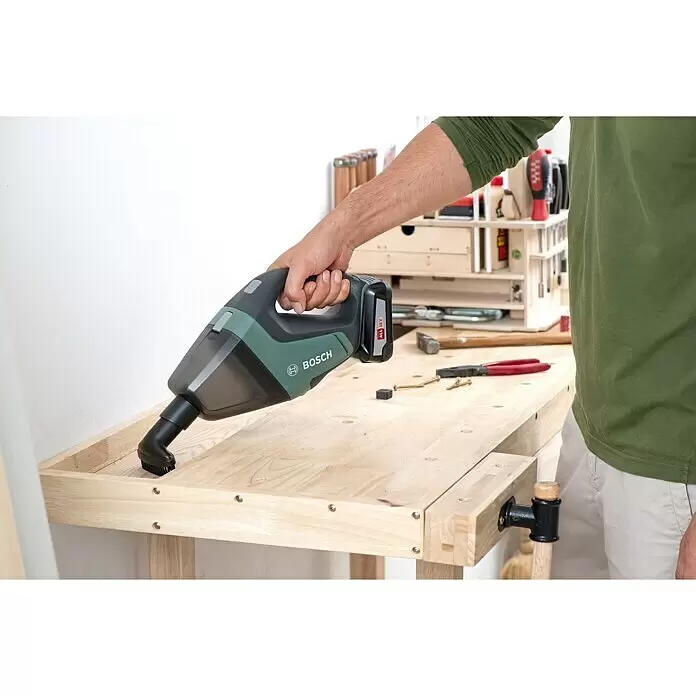 Bosch Power for All 18V Aspirador de mano de batería UniversalVac 18 - 18 V, Sin batería, Volumen de recipiente: 0,5 l - Una persona utiliza un aspirador de mano Bosch verde y negro con cepillo para limpiar un banco de madera.