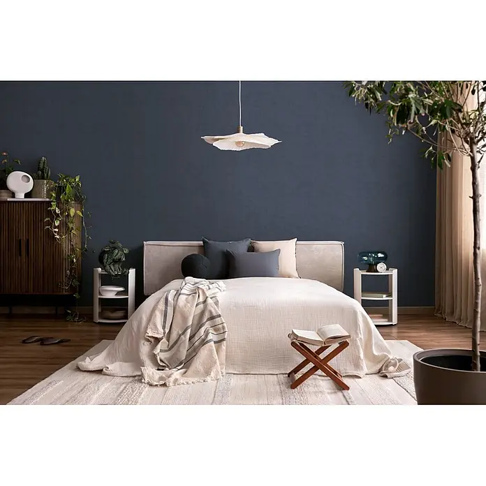 A.S. Création Nordic Feeling Vliestapete Uni - Blau, Uni, 10,05 x 0,53 m - Ein gemütliches Schlafzimmer mit einem Bett, hellcremefarbener Bettwäsche, verschiedenen Kissen und einer gestreiften Decke, vor einer dunkelblauen Wand.