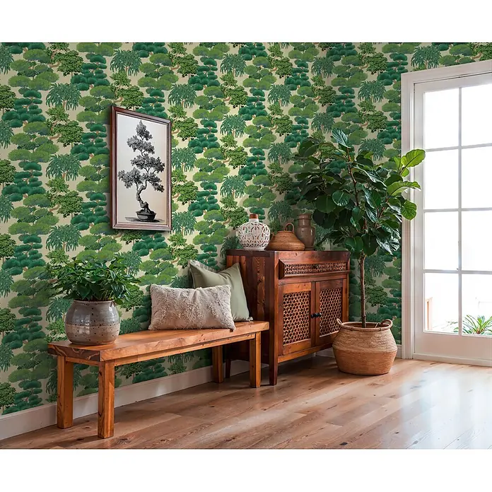 A.S. Création Cuba 2 Vliestapete Natur - Grün-Creme, Floral, 10,05 x 0,53 m - Grüne Tapete mit dichtem Baummuster bedeckt eine Wand und schafft eine üppige, natürliche Atmosphäre in einem Raum mit Holzmöbeln und Pflanzen.