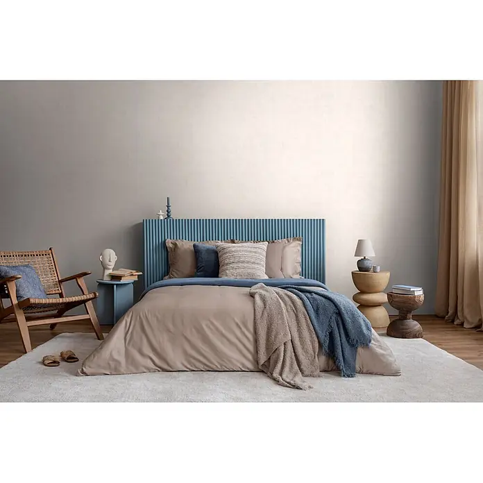 A.S. Création Nordic Feeling Vliestapete Uni - Grau-Weiß, Uni, 10,05 x 0,53 m - Modernes Schlafzimmer mit Bett, beiger Bettdecke, blauen und beigen Überwürfen und markantem blauem geriffeltem Kopfteil auf hellem Teppich.