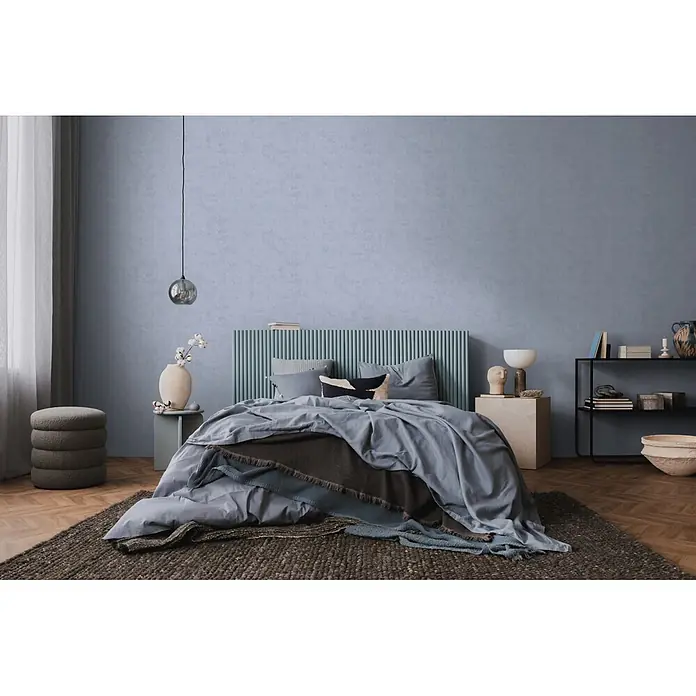 A.S. Création Nordic Feeling Vliestapete Uni - Blau-Grau, Uni, 10,05 x 0,53 m - Ein modernes Schlafzimmer mit einem Bett mit blau-grauer Bettdecke, mehreren Überwürfen und einem grünen Lattenkopfteil.