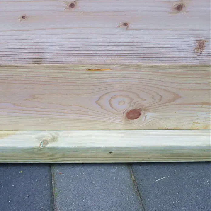 Alpholz Gartenhaus Universal Holzfußboden-Set 30 m² - Naturbelassen - Helle Holzbretter mit Naturmaserung, darunter ein behandeltes Brett, horizontal über einem gepflasterten Untergrund gestapelt.