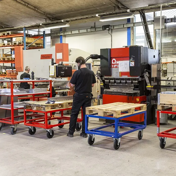 Kongamek Palettenwagen Palettenwagen in rot 650mm hoch mit Bremse für Paletten 1200x1000mm -  - Arbeiter bedienen industrielle Abkantpressen in einer Metallverarbeitungswerkstatt, umgeben von Transportwagen und Paletten.