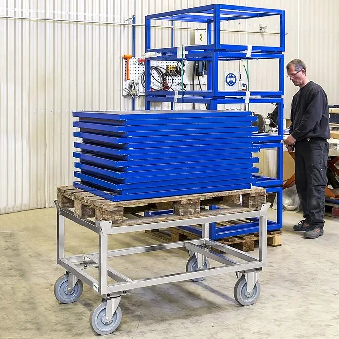 Kongamek Palettenwagen Palettenwagen in rot 305mm hoch ohne Bremse für Euro-Paletten 1200x800mm -  - Silberner Metallwagen mit Rollen trägt eine Holzpalette mit gestapelten blauen Rahmen in einer Werkstatt.
