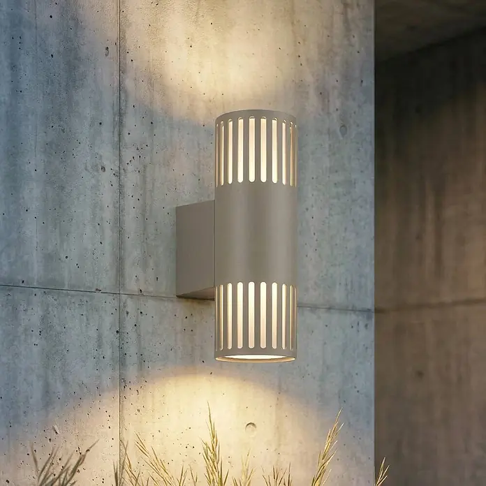 Lindby Wandleuchte aussen in grau, weiß, Aussenlampe Wand -  - Graue zylindrische Außenwandleuchte mit vertikalen Schlitzen, leuchtet auf eine Betonwand, mit trockenem Gras unten.