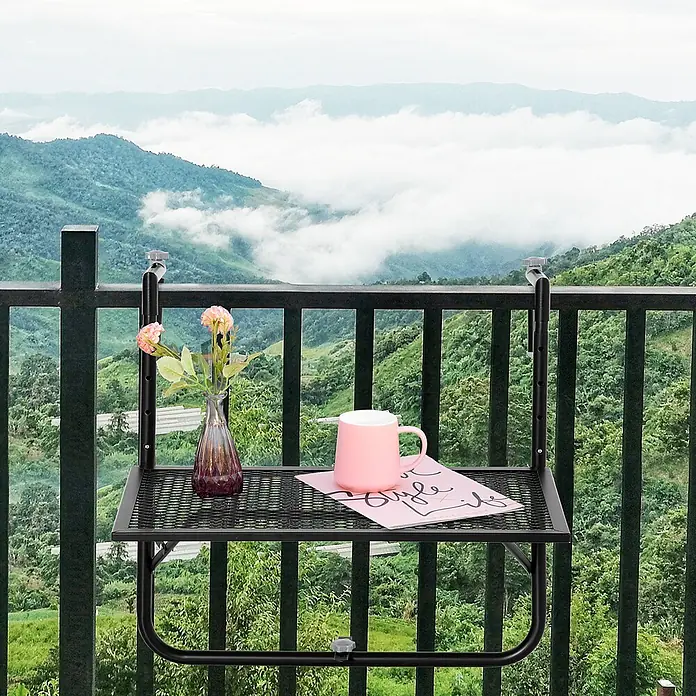 Outsunny Balkon-Hängetisch - Schwarz - Schwarzer Metall-Balkontisch mit Gitterfläche, auf dem eine rosa Tasse, eine Vase mit Blumen und ein Buch stehen, an einem Geländer mit Bergblick.